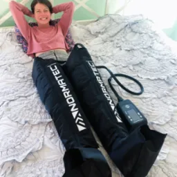 Uśmiechnięta kobieta leży na łóżku z nałożonymi na nogi czarnymi mankietami kompresyjnymi marki Normatec, podłączonymi do panelu sterowania.