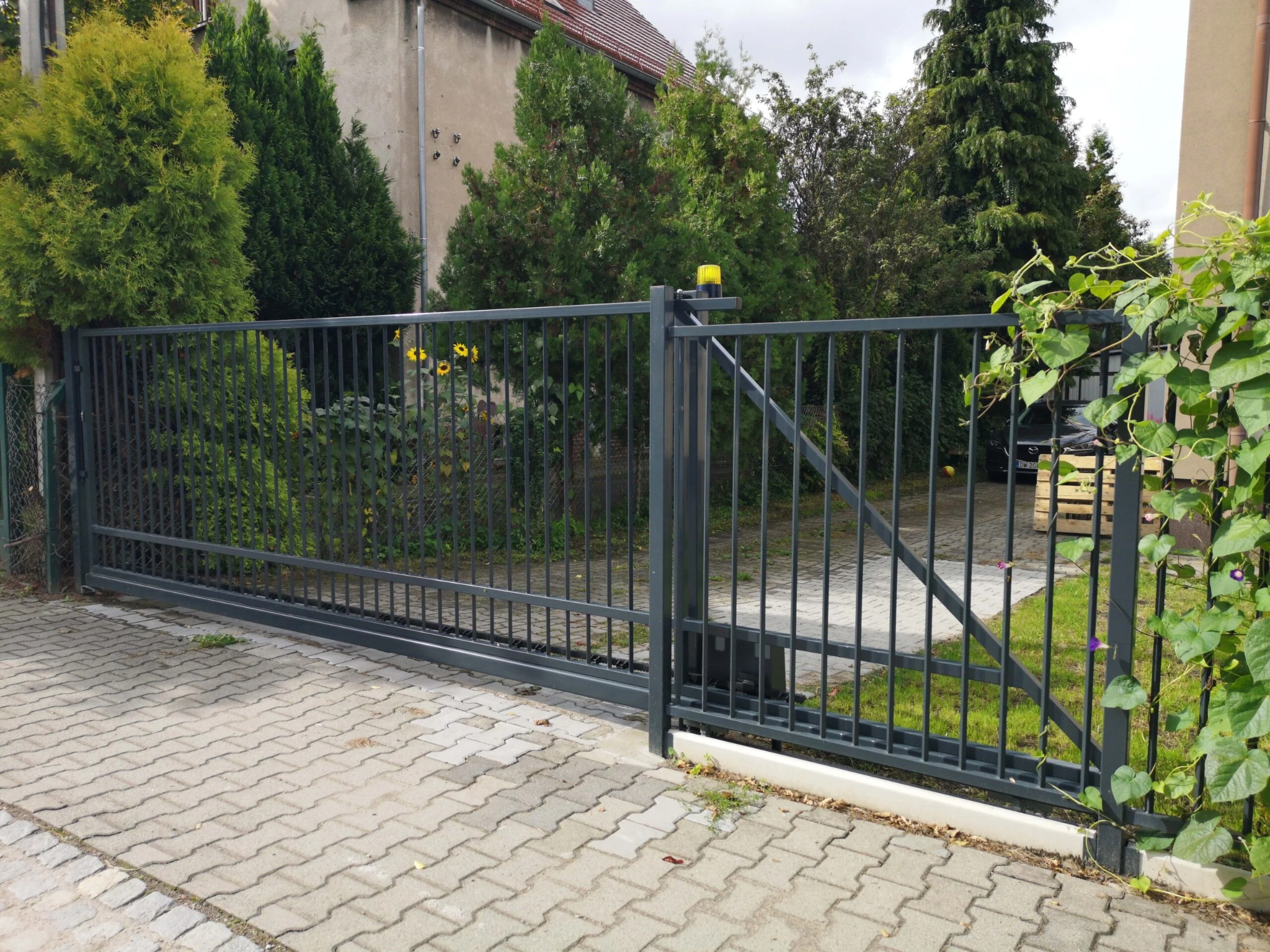 Przesuwna brama wjazdowa z metalowymi prętami, szara, z żółtą lampą ostrzegawczą, widok z zewnątrz na kostkę brukową i zieleń ogrodu.
