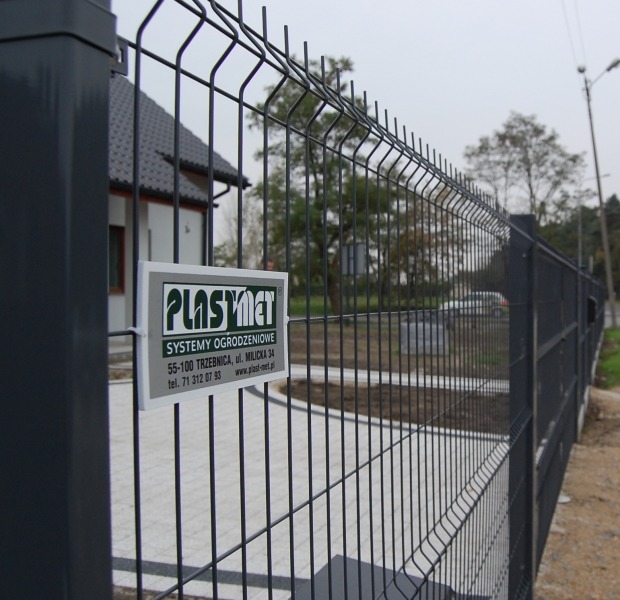 Grafitowe ogrodzenie panelowe z tabliczką informacyjną producenta, widok z bliska na słupku, w tle dom i kostka brukowa.