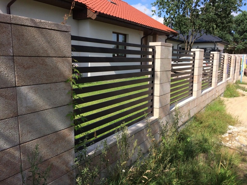 Nowoczesne ogrodzenie z poziomych, czarnych profili między słupkami z szarej kostki, widok z bliska na tle zieleni i domów.