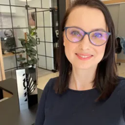 Portret uśmiechniętej kobiety w okularach na tle biura z przeszklonymi ścianami i rośliną doniczkową, obok stoi nagroda.