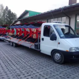 Biały bus z przyczepą przewozi pługi rolnicze na tle budynku w Krośnie. Pługi są w kolorze czerwonym i czarnym.