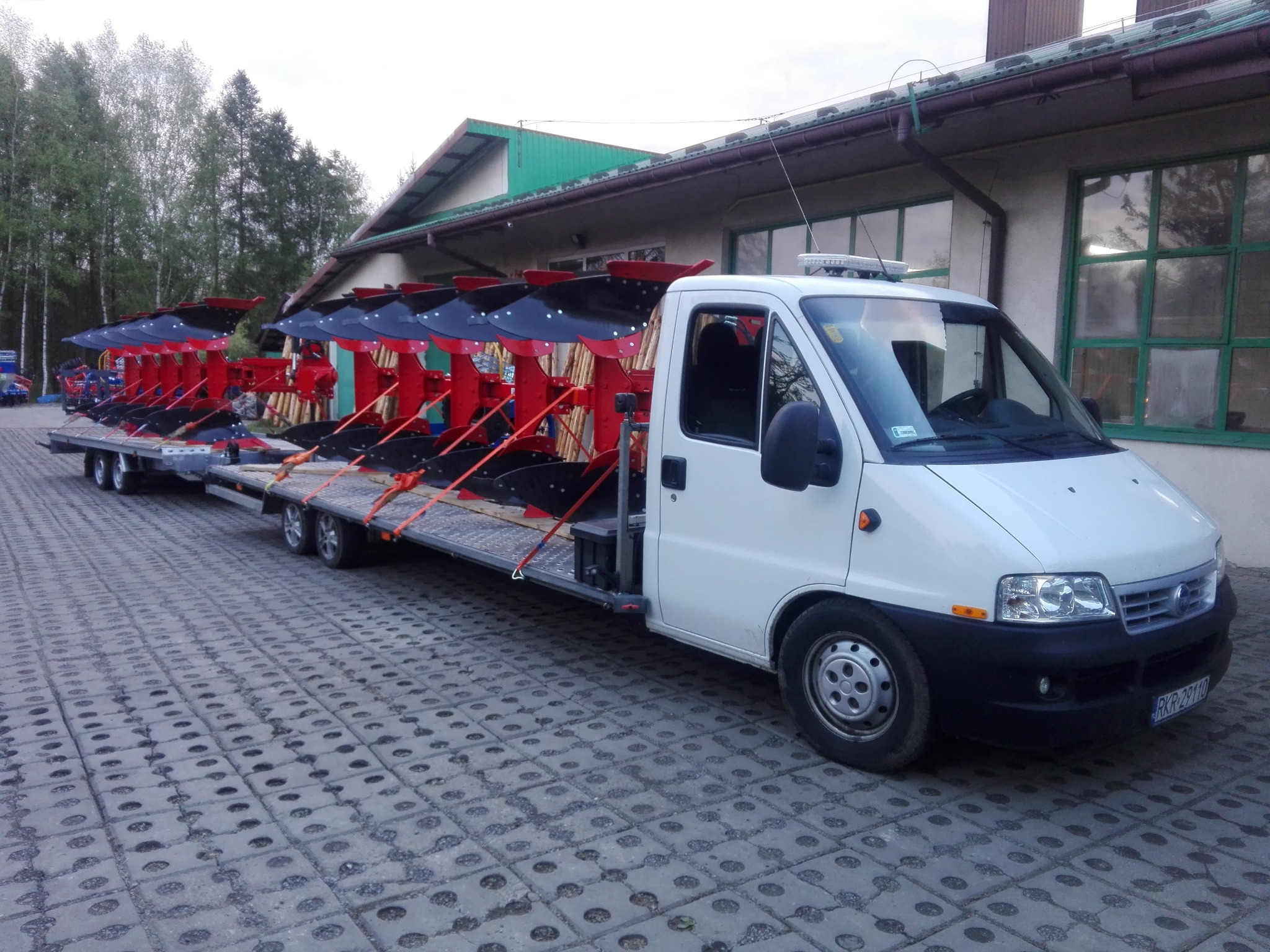 Biały bus z przyczepą przewozi pługi rolnicze na tle budynku w Krośnie. Pługi są w kolorze czerwonym i czarnym.