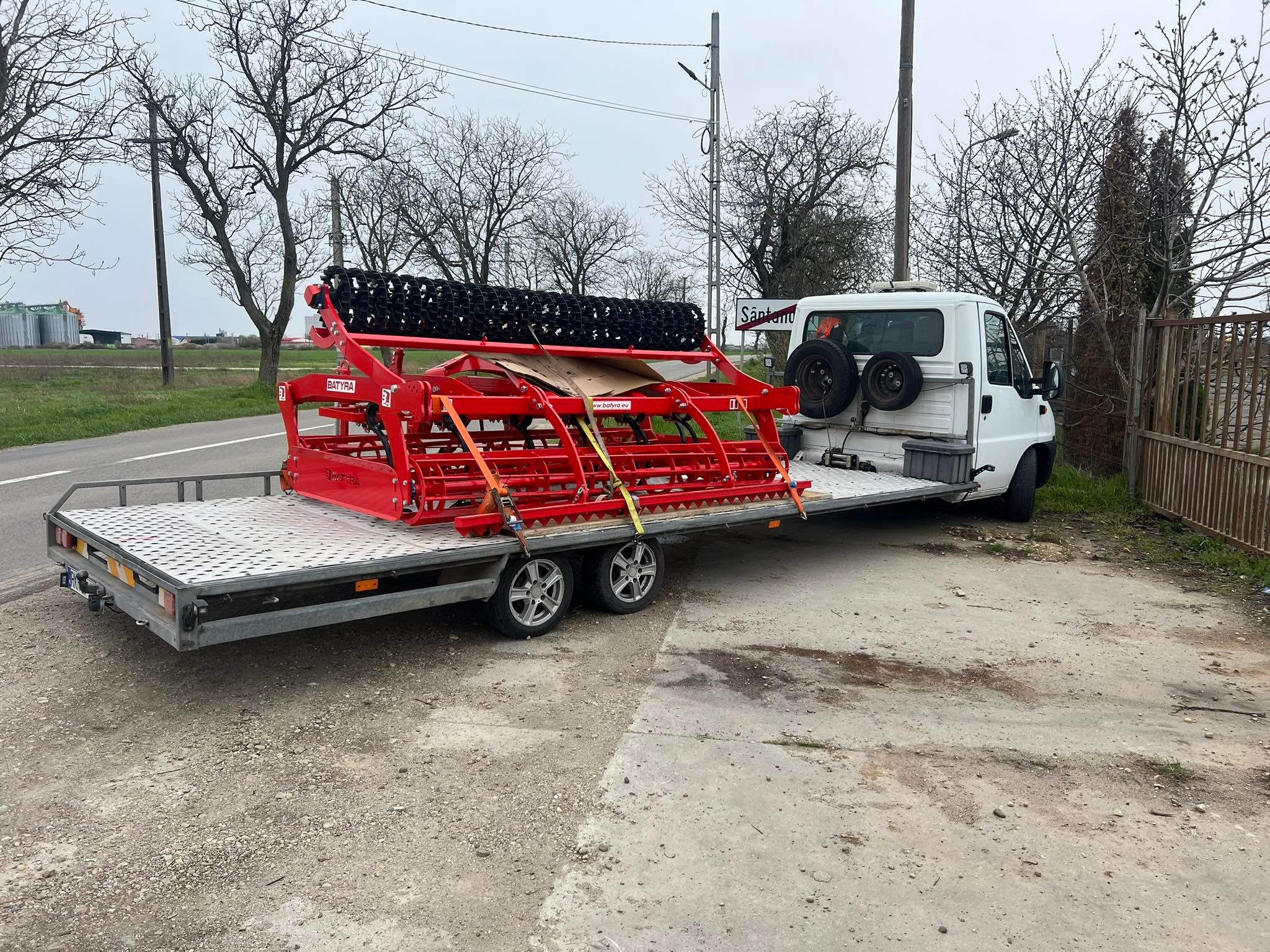 Biały bus z przyczepą przewożący czerwony sprzęt rolniczy marki Batyra, zabezpieczony pasami, na tle szarej drogi i drzew bez liści.