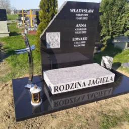 Czarny nagrobek z granitu z wygrawerowanymi imionami i datami, ozdobiony srebrnym krzyżem i lampionem, na tle cmentarza z zielenią i innymi nagrobkami.