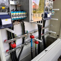 INSTAL3D Shevchenko Dmytro - Otwarta skrzynka elektryczna z widocznymi przewodami, bezpiecznikami, listwami zaciskowymi i elementami montażowymi, z zewnątrz widoczny fragment budynku.