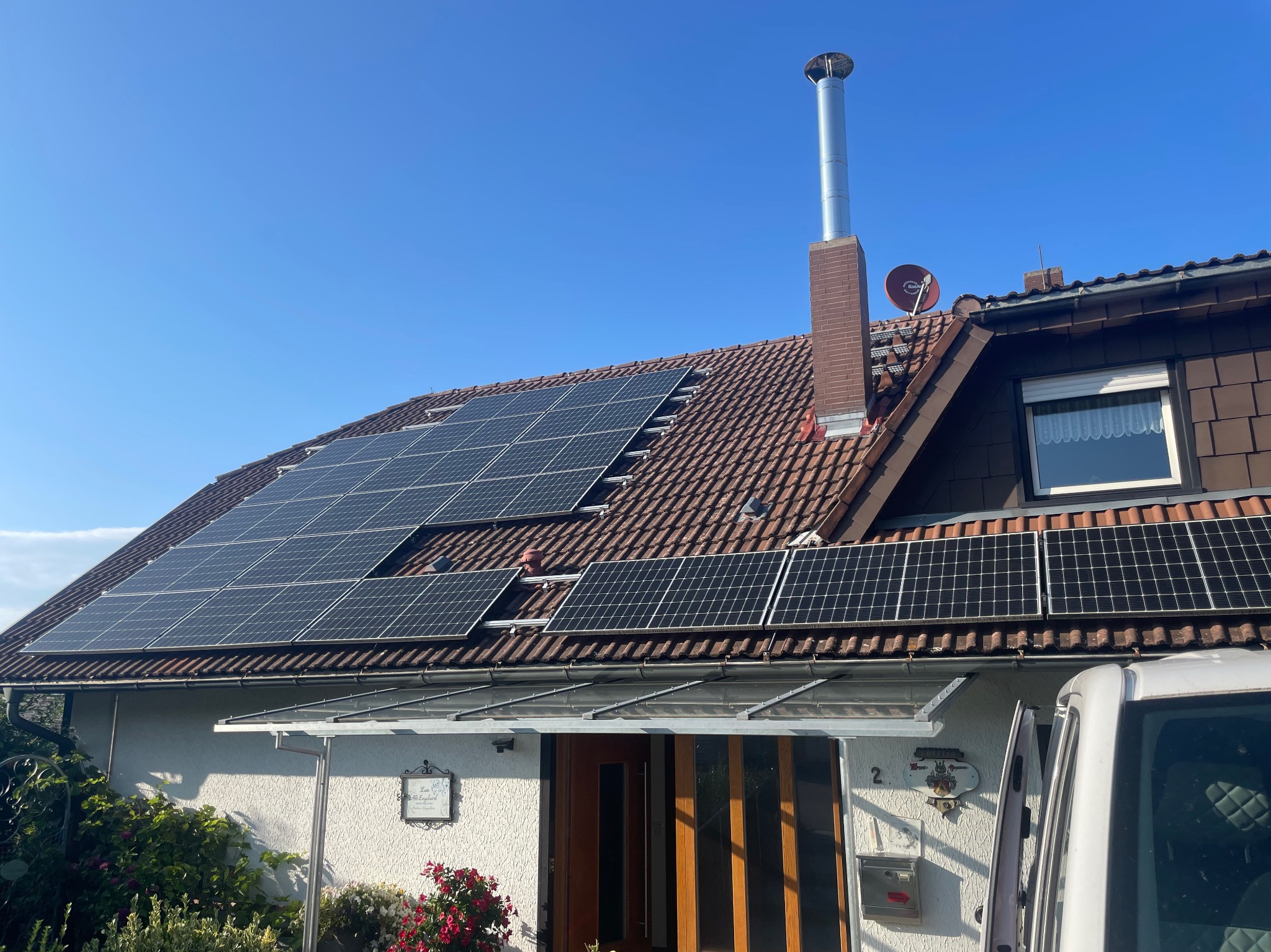 Dom jednorodzinny z nowo zainstalowanymi panelami słonecznymi na brązowym dachu, widoczny komin z metalowym zakończeniem, antena satelitarna i fragment samochodu dostawczego z otwartymi drzwiami.