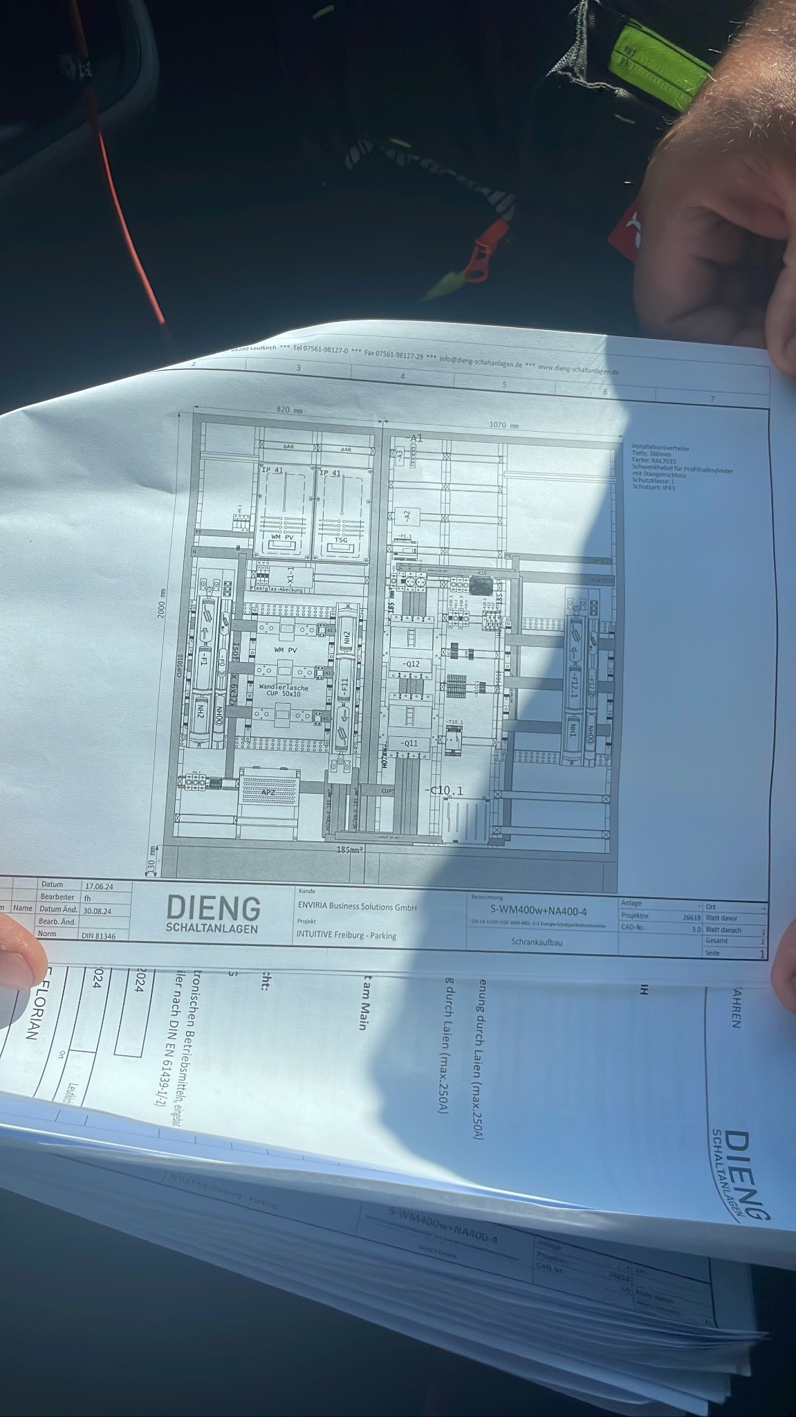 Schemat elektryczny rozdzielnicy w formacie A3, z naniesionymi wymiarami i oznaczeniami komponentów, trzymany w ręku, widoczne fragmenty instalacji elektrycznej i oznaczenia producenta.