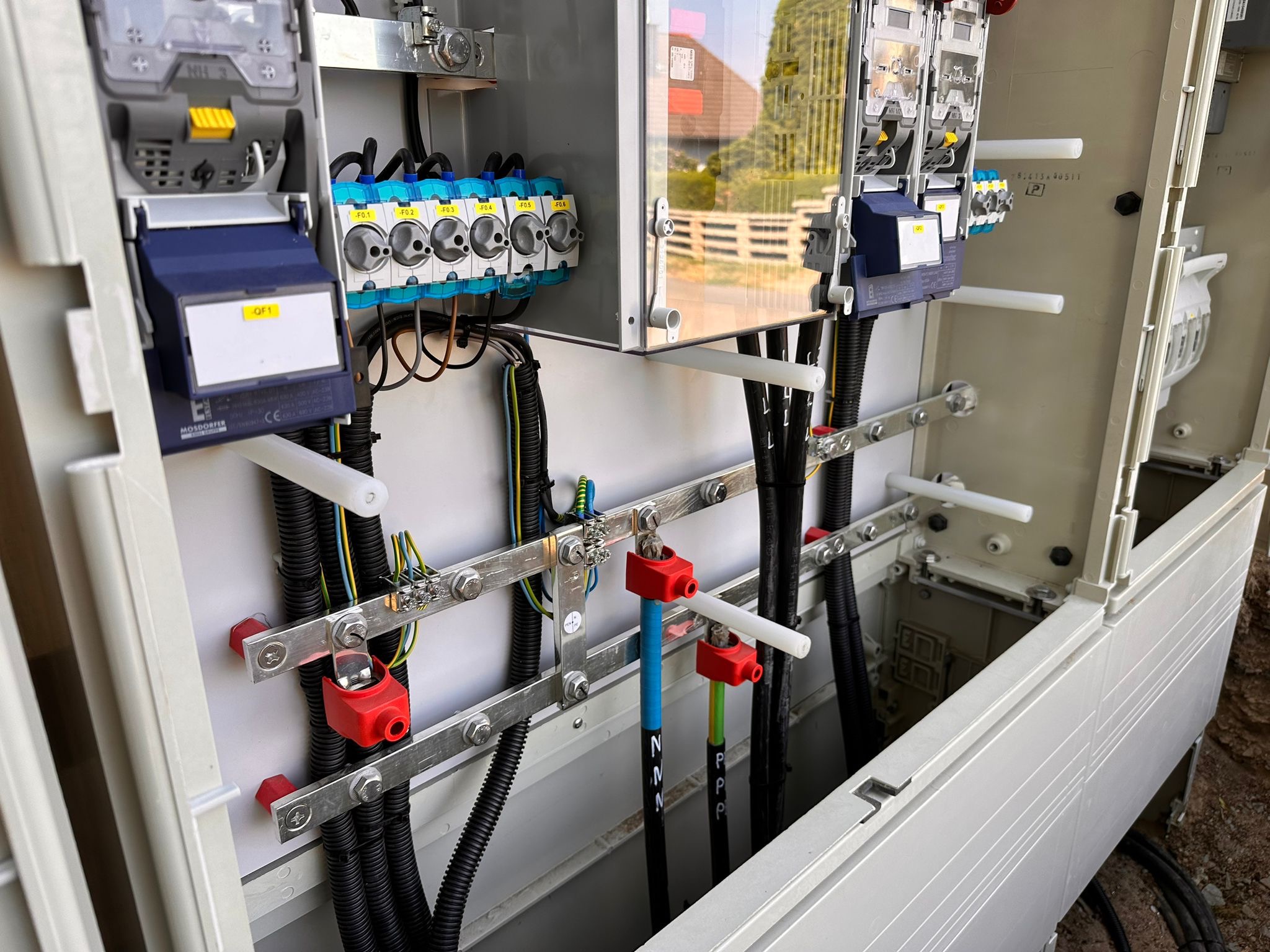 Otwarta skrzynka elektryczna z widocznymi przewodami, bezpiecznikami, listwami zaciskowymi i elementami montażowymi, z zewnątrz widoczny fragment budynku.