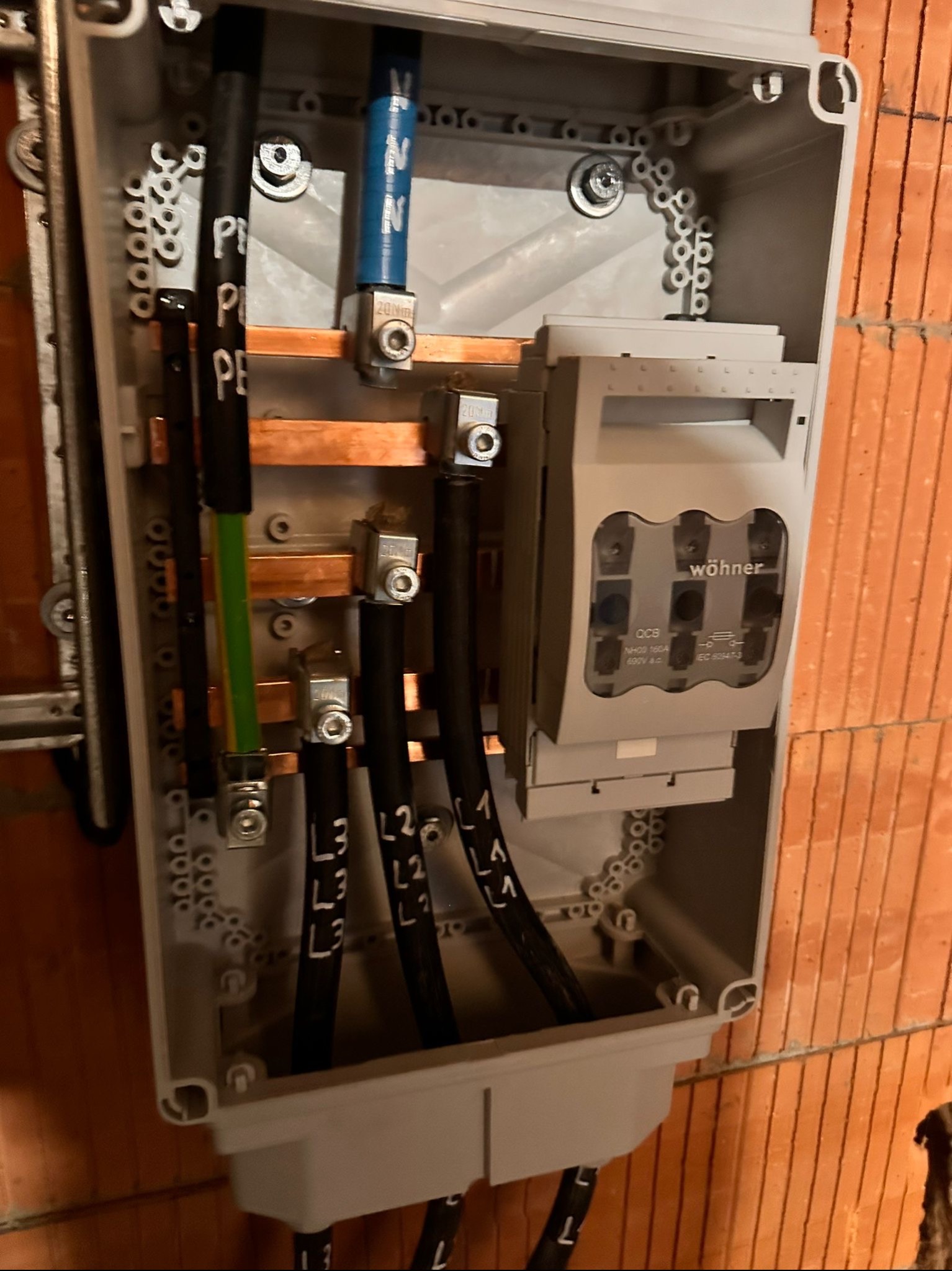 Rozdzielnica elektryczna w trakcie montażu, widoczne kable z oznaczeniami L1, L2, L3 oraz PE, szyny miedziane i bezpieczniki firmy Wöhner, zamontowana na ścianie z cegły.