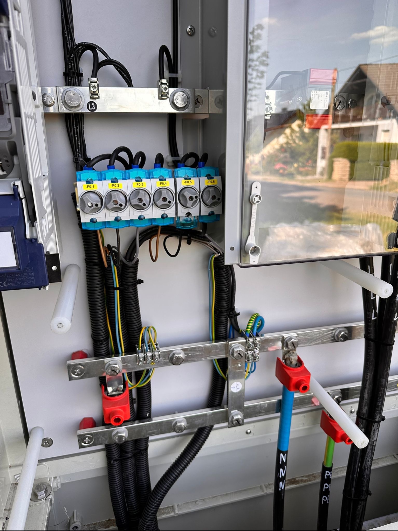 Wnętrze skrzynki elektrycznej z bezpiecznikami, okablowaniem w peszlach i listwami uziemiającymi, widoczne oznaczenia przewodów N i PE.