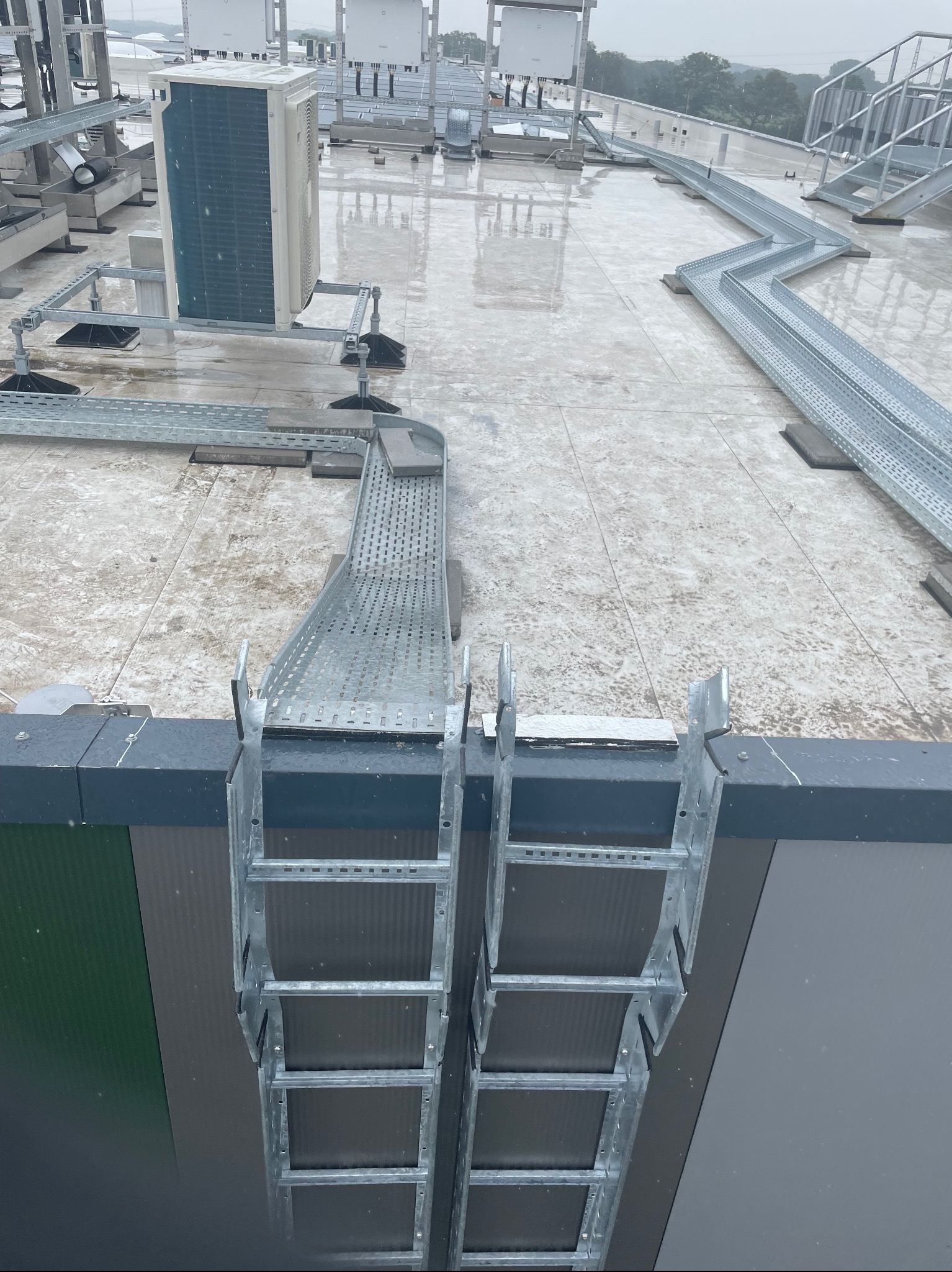 Instalacja przemysłowa na dachu: koryta kablowe, klimatyzator i drabina dostępowa w deszczowy dzień.