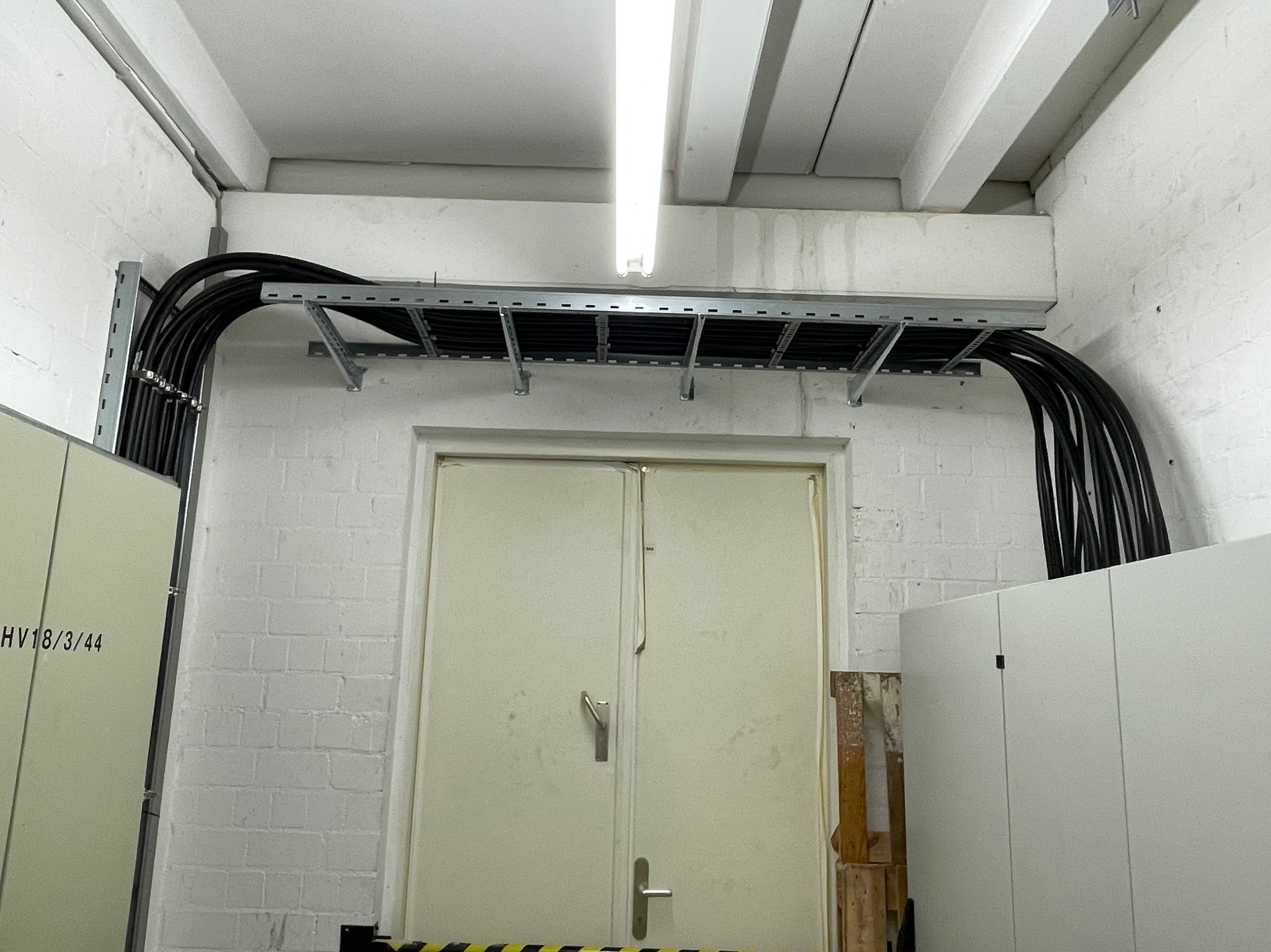 Czarny kabel elektryczny ułożony w korytkach na ścianie z białej cegły nad białymi drzwiami w pomieszczeniu technicznym.