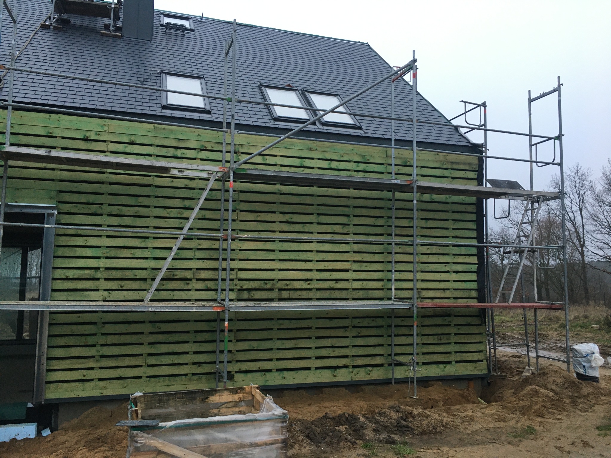 Dom w trakcie budowy z szarym dachem łupkowym i zieloną drewnianą elewacją, widoczne rusztowania i okna dachowe.