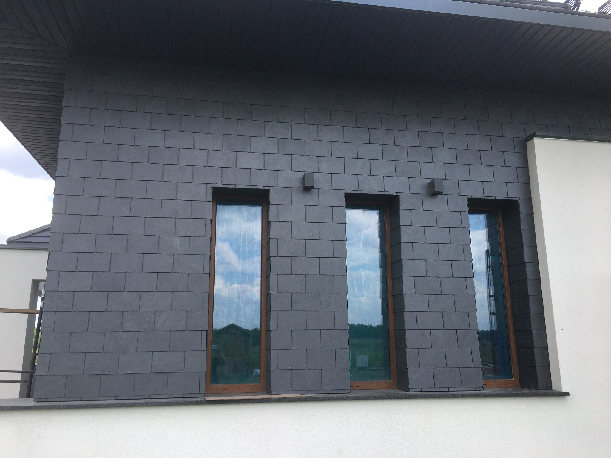 Elewacja domu wykończona szarym, łupkowym kamieniem elewacyjnym ułożonym w cegiełkę, z trzema oknami o drewnianych ramach, w których odbija się błękitne niebo i zieleń drzew.