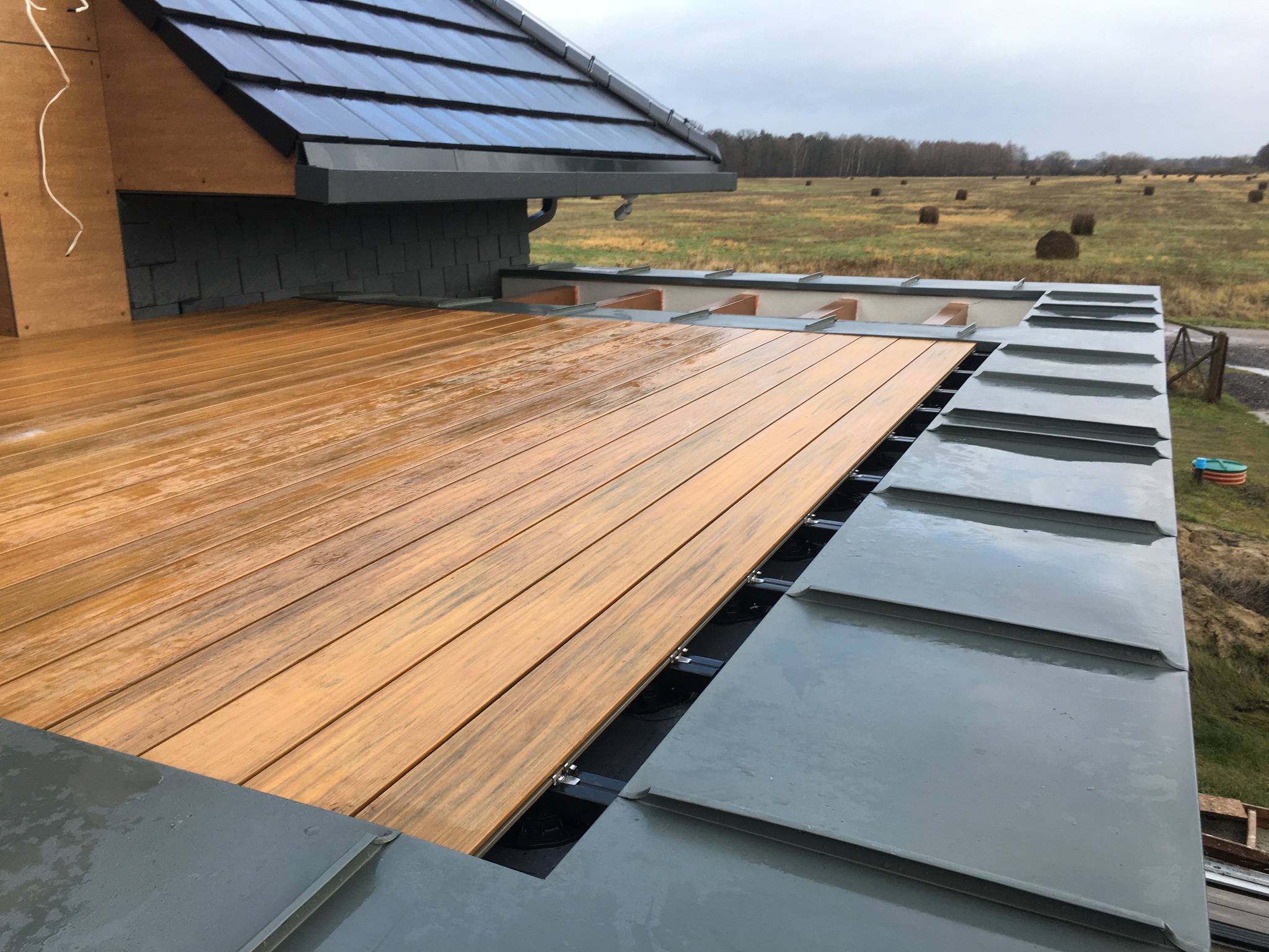 Nowoczesny dach z szarej blachy trapezowej połączony z drewnianym tarasem, widok na pole z balotami siana w tle, pochmurne niebo.