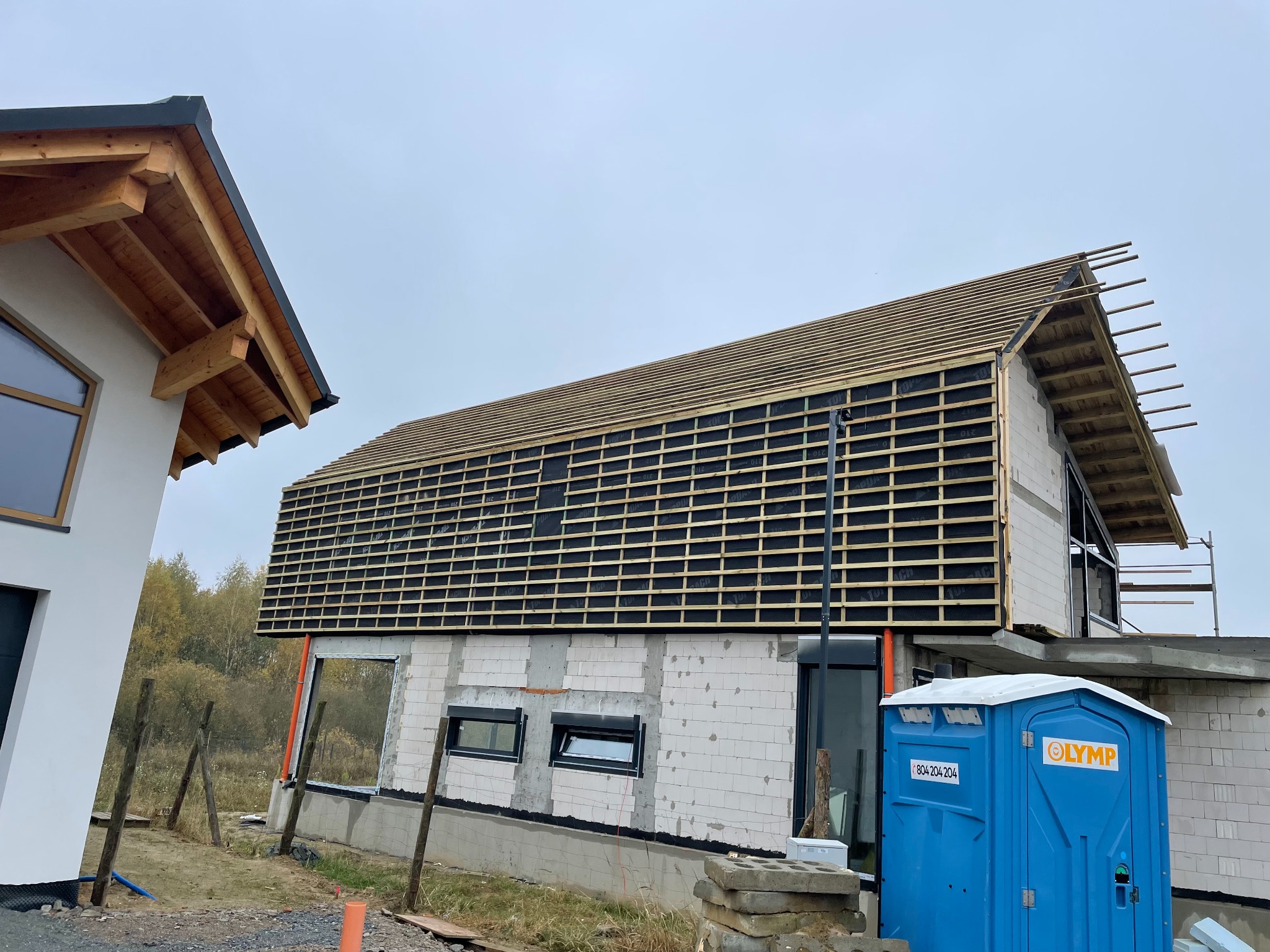 Budowa domu jednorodzinnego: widok na konstrukcję dachu z łatami i membraną, obok drugi budynek z widocznym fragmentem dachu, na pierwszym planie przenośna toaleta.