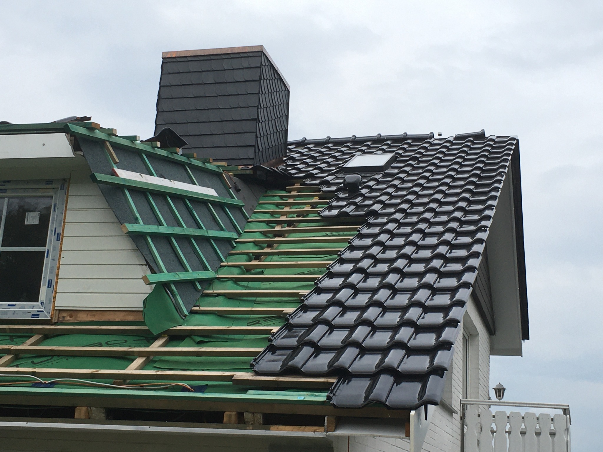 Fragment dachu w trakcie prac: część pokryta błyszczącą, czarną dachówką, druga część w trakcie przygotowania z widoczną membraną, łatami i kontrłatami. Widoczny komin obłożony łupkiem.