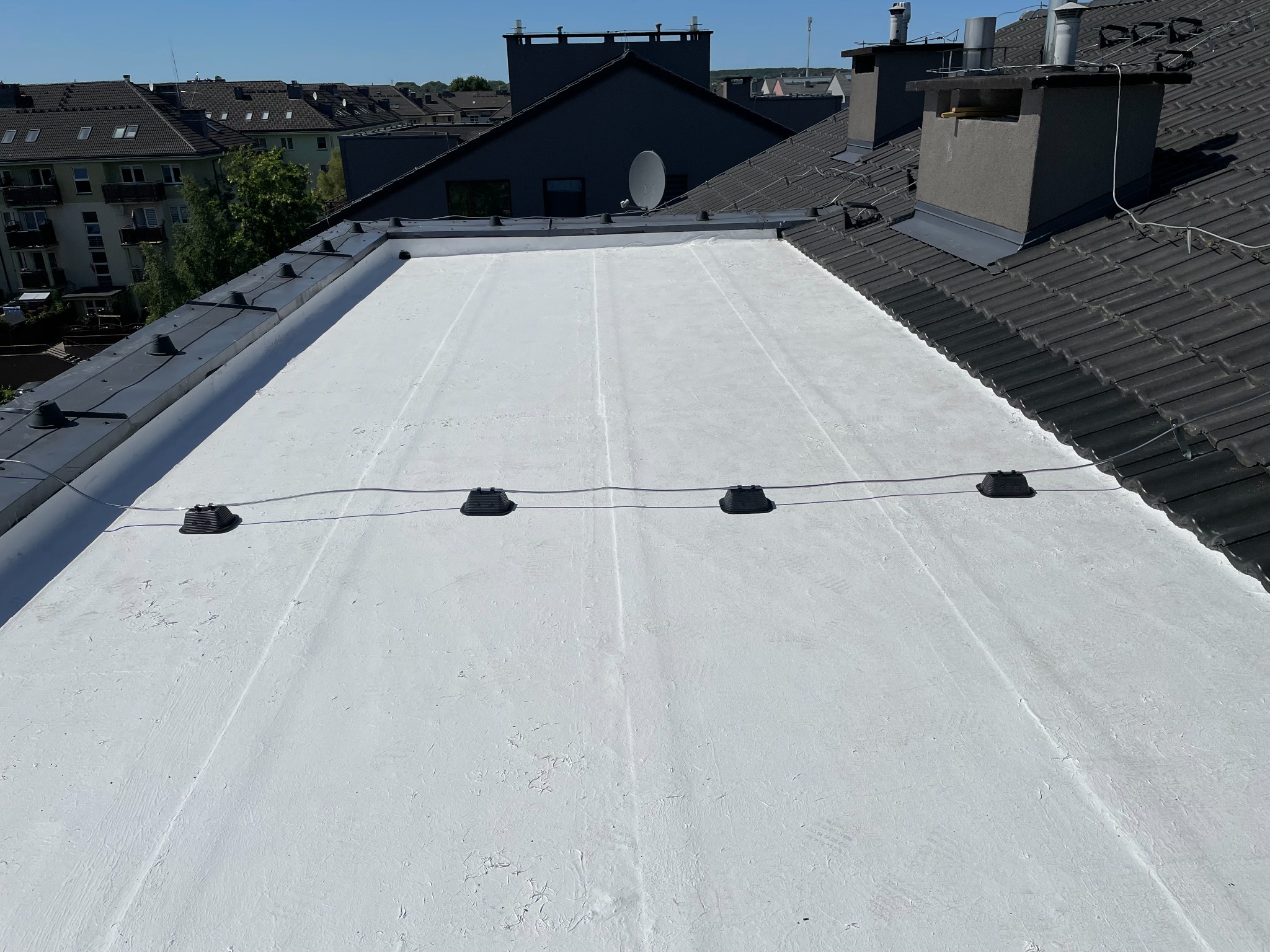 Biała, płaska powierzchnia dachu z widocznymi elementami instalacji odgromowej i fragmentami sąsiednich dachów pokrytych ciemną dachówką w tle.
