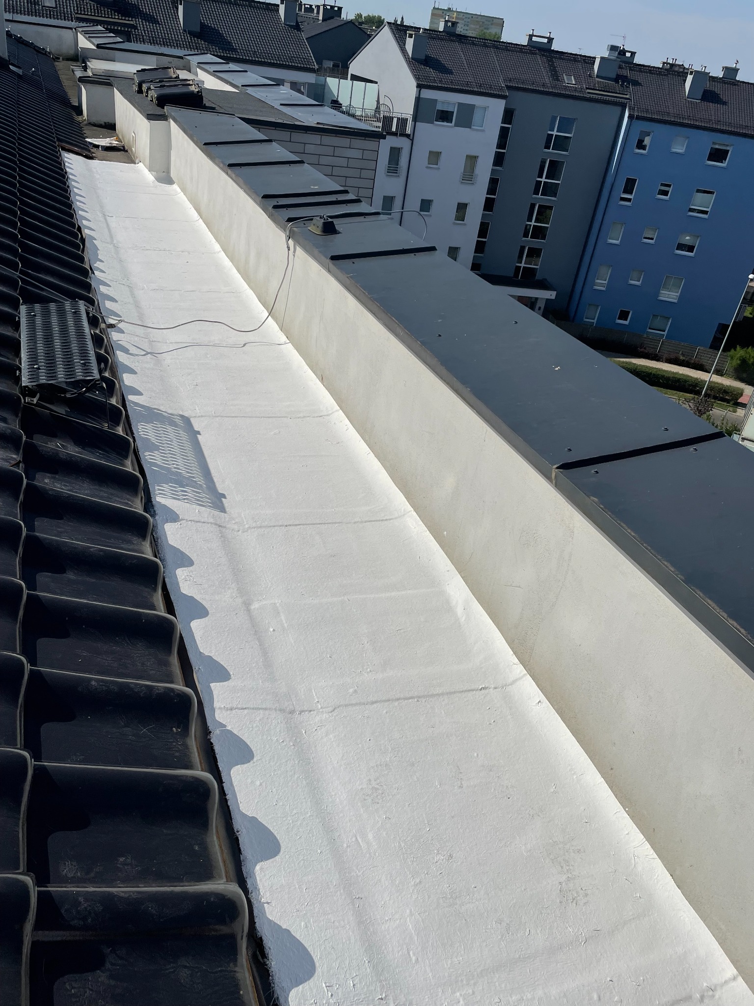 Widok z góry na płaski dach pokryty białą hydroizolacją, sąsiadujący z dachem skośnym pokrytym czarną dachówką, w tle budynki mieszkalne.
