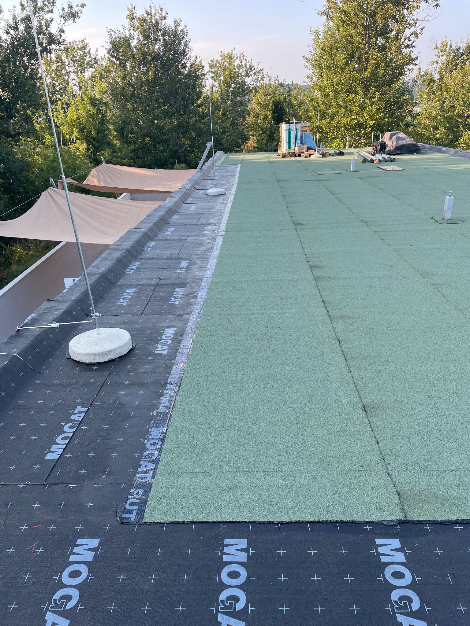 Płaski dach pokryty zieloną membraną bitumiczną, częściowo wykończony czarną membraną z białym nadrukiem 'MOGAT', widoczne elementy instalacji odgromowej, w tle zieleń drzew.