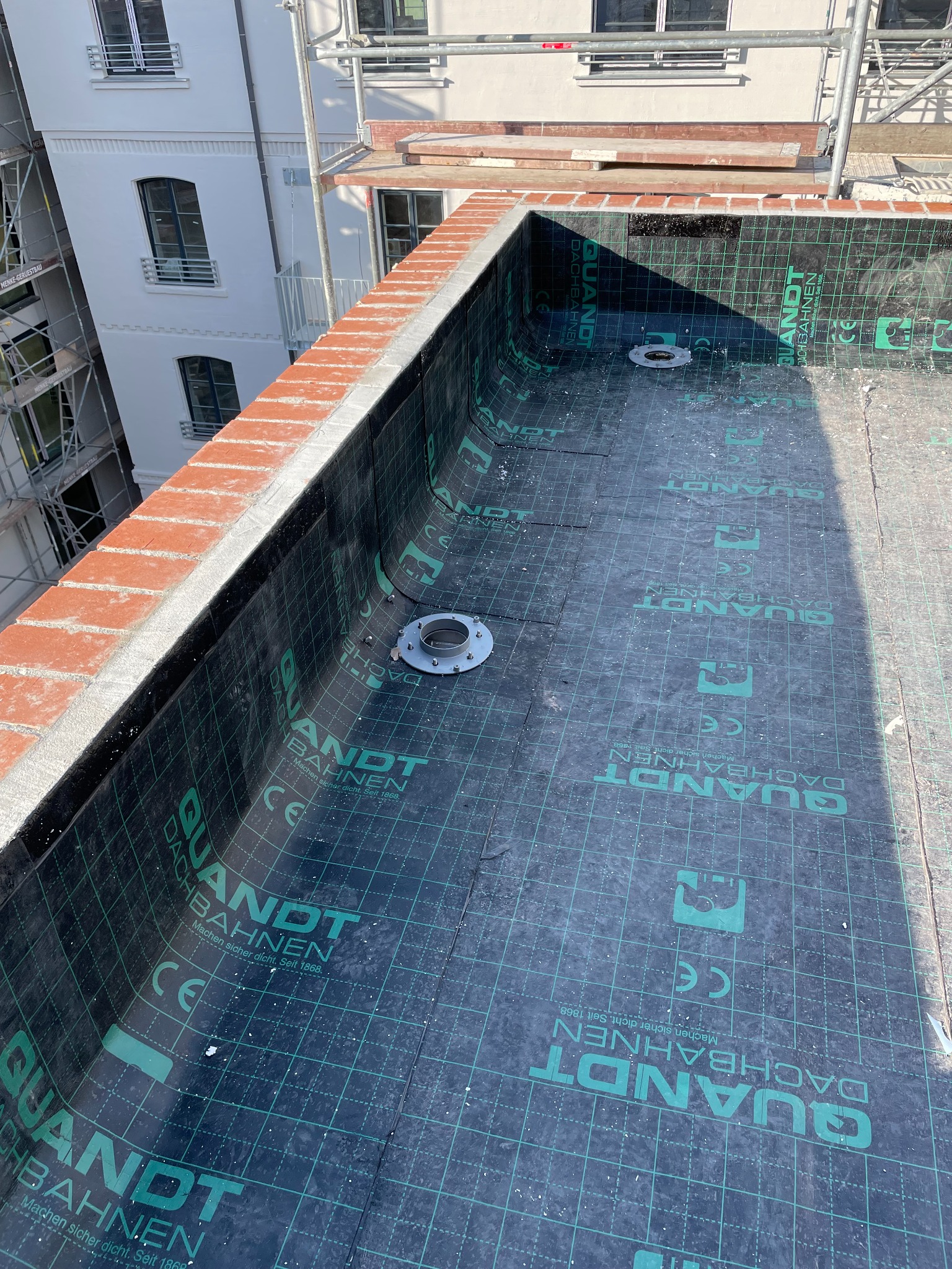 Widok z góry na płaski dach z hydroizolacją z papy z logo QUANDT DACHBAHNEN, obróbki blacharskie przy krawędzi dachu z ceglanym wykończeniem, w tle budynek w rusztowaniach.