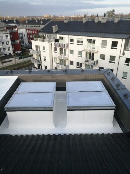 Dwa świetliki dachowe na czarnym dachu z dachówki ceramicznej, uszczelnione białą masą, w tle budynki mieszkalne.