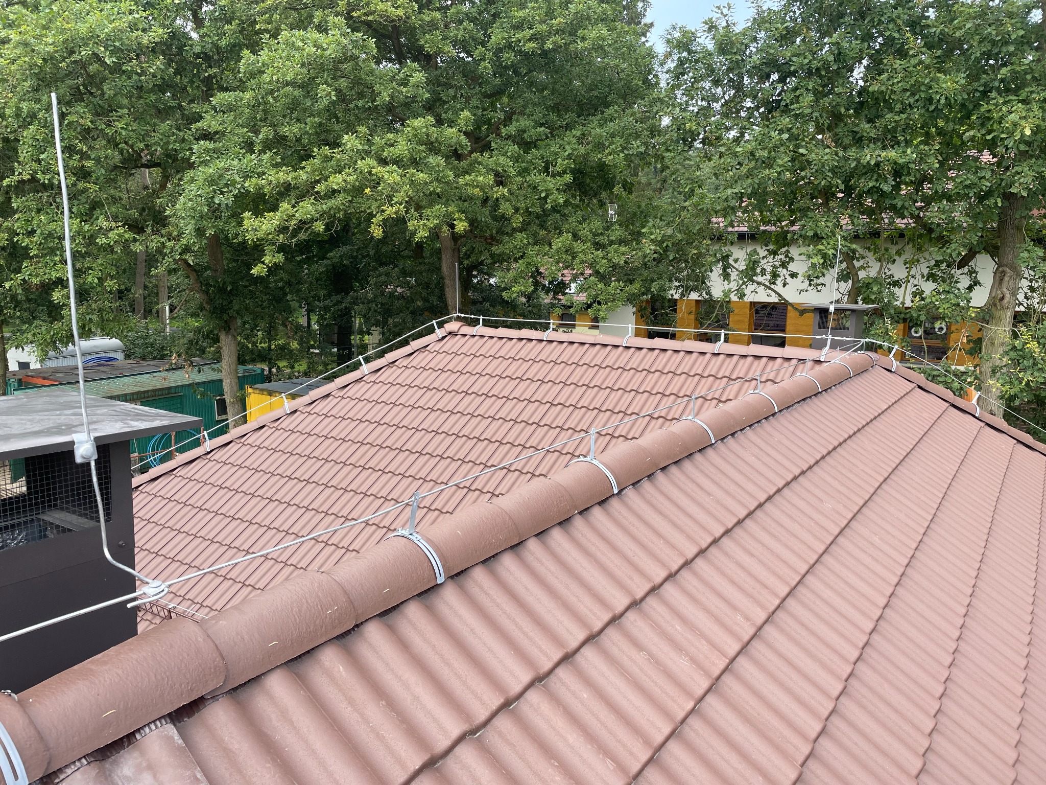 Czerwono-brązowy dach z dachówki ceramicznej, zabezpieczony systemem odgromowym z poziomymi przewodami mocowanymi metalowymi uchwytami, na tle zielonych drzew.