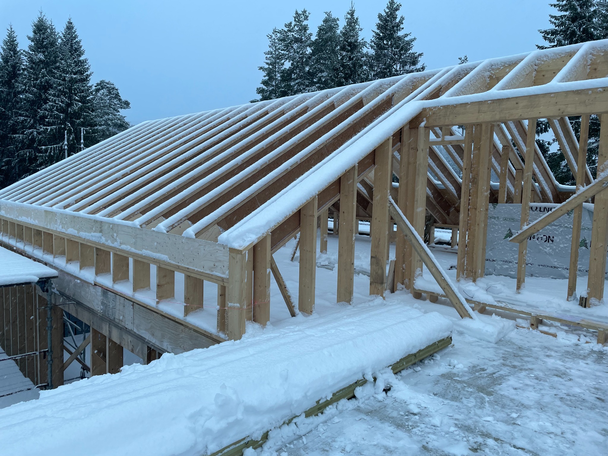 Drewniana konstrukcja dachu w trakcie budowy, pokryta śniegiem, z widocznymi drzewami w tle w pochmurny dzień.