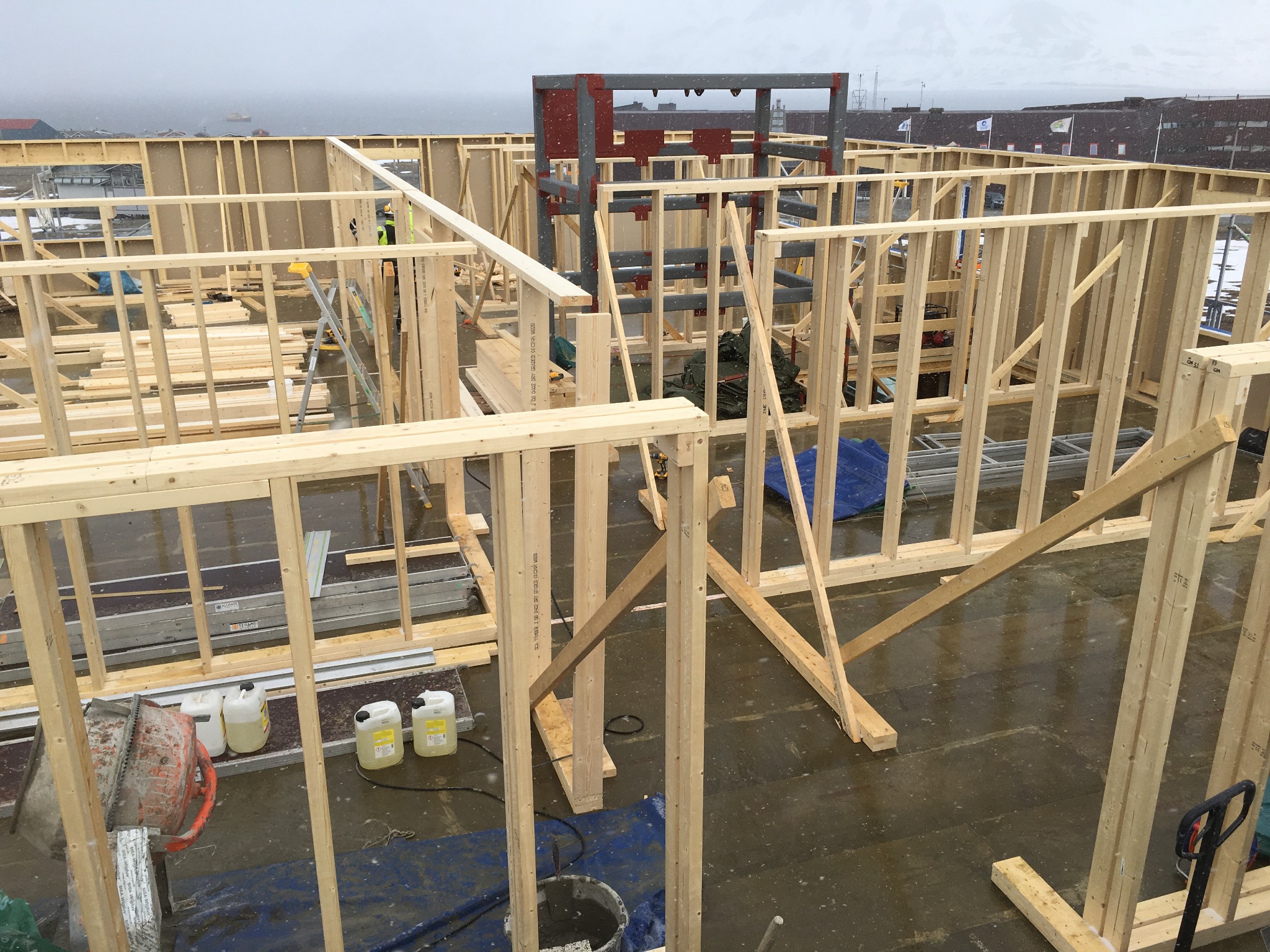 Konstrukcja drewniana dachu w trakcie budowy, widok z góry na szkielet z belek, w tle widać zabudowania i mglisty krajobraz, na podłożu leżą narzędzia budowlane i pojemniki z płynami.