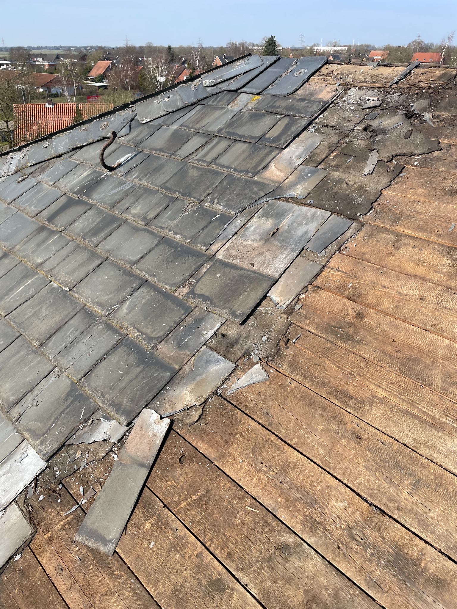 Uszkodzony dach z szarej dachówki łupkowej, z widocznymi drewnianymi deskami poszycia. Fragmenty dachówek rozsypane na powierzchni. W tle zabudowa miejska.