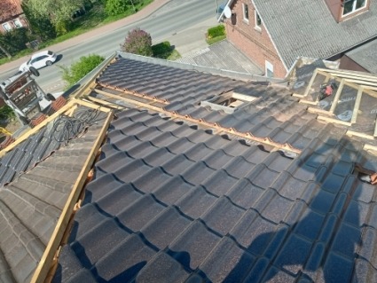 Częściowo pokryty dach z ciemnej blachodachówki, widać elementy konstrukcji drewnianej i niezakończone fragmenty. W tle budynki i ulica.