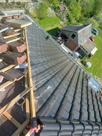 Częściowy widok dachu w trakcie układania ciemnej dachówki, z widocznym drewnianym szkieletem i stosem dachówek. W tle zieleń i zabudowania gospodarcze.