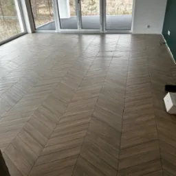 Ułożona w jodełkę podłoga z płytek imitujących drewno w przestronnym, nowoczesnym wnętrzu z dużymi oknami balkonowymi i fragmentem ciemnozielonej ściany.