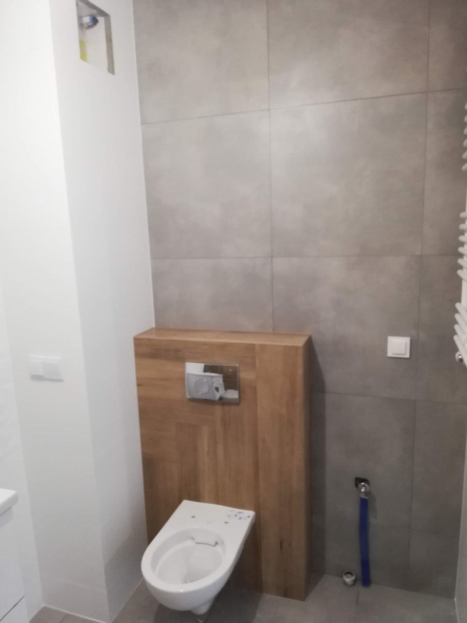 Nowoczesna łazienka z szarymi płytkami, zabudową WC z drewnopodobnej płyty i chromowanym przyciskiem spłukiwania.