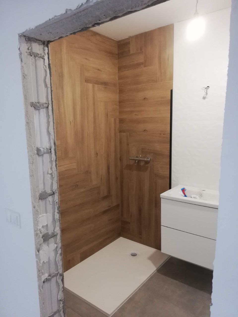 Remontowana łazienka z kabiną prysznicową wyłożoną płytkami imitującymi drewno w jodełkę, białą umywalką z szafką i niedokończonym ościeżem drzwi.