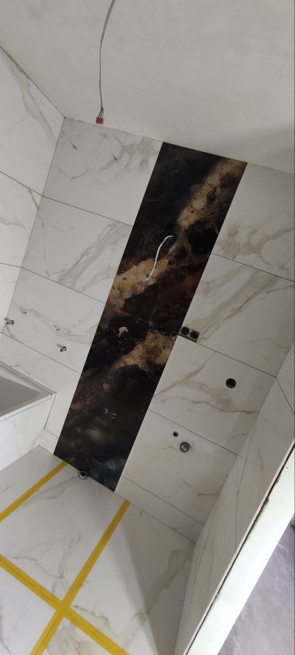 Wnętrze łazienki w trakcie prac wykończeniowych: płytki marmurowe na ścianach i podłodze, ciemny pas dekoracyjny na ścianie, wystające kable, żółta taśma zabezpieczająca na podłodze, widoczne...