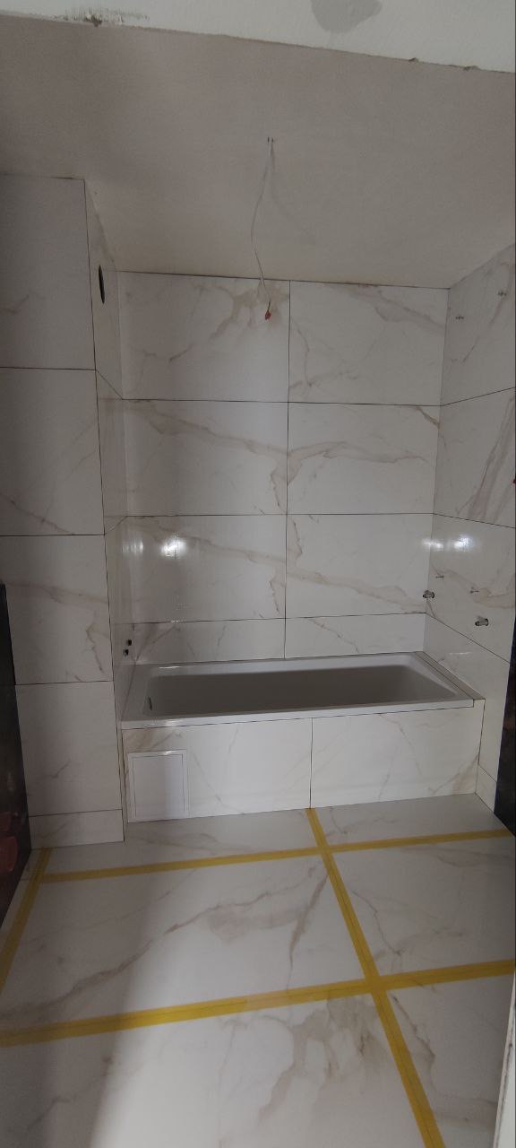 Wnętrze łazienki w trakcie remontu, z białą wanną obudowaną płytkami imitującymi marmur, widoczne zabezpieczenia podłogi taśmą malarską, surowy sufit z wystającymi przewodami.