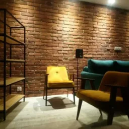 Salon z dekoracyjną ścianą z cegły, oświetlony punktowo, z metalowym regałem z drewnianymi półkami, dwoma fotelami w odcieniach żółci i turkusową kanapą.