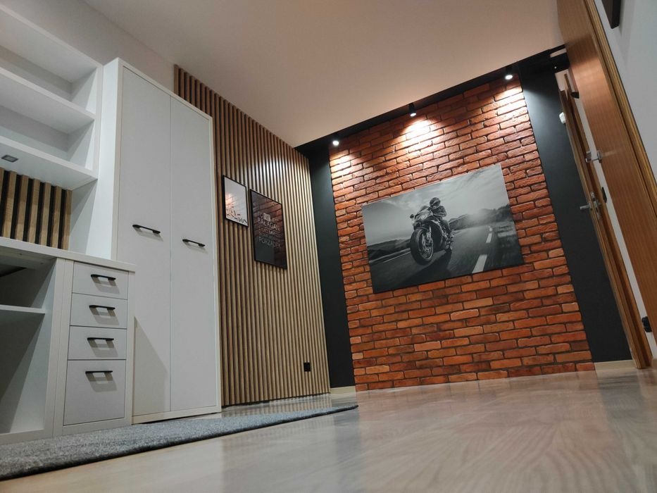Nowoczesne wnętrze z dekoracyjną ścianą z cegły, na której wisi czarno-biały obraz motocykla, obok ściana z drewnianych listewek, białe meble biurowe i szara wykładzina dywanowa.