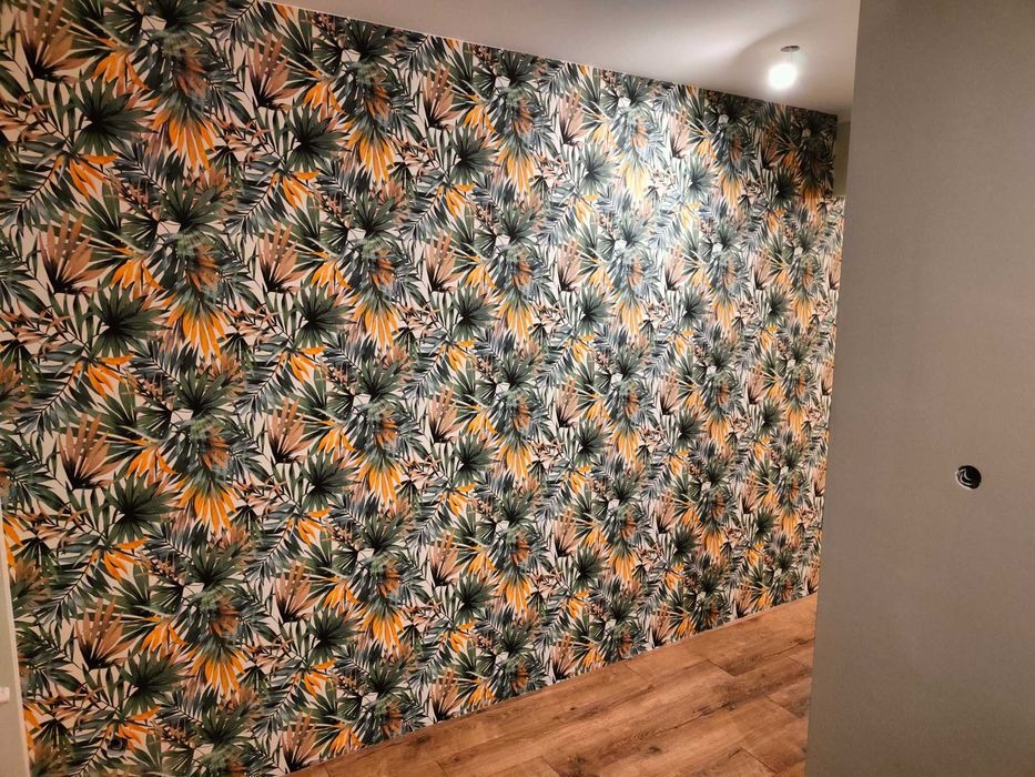 Ściana pokryta tapetą z motywem tropikalnych liści w odcieniach zieleni i pomarańczy, widoczna z perspektywy podłogi wyłożonej panelami imitującymi drewno, obok ściana pomalowana na szaro...