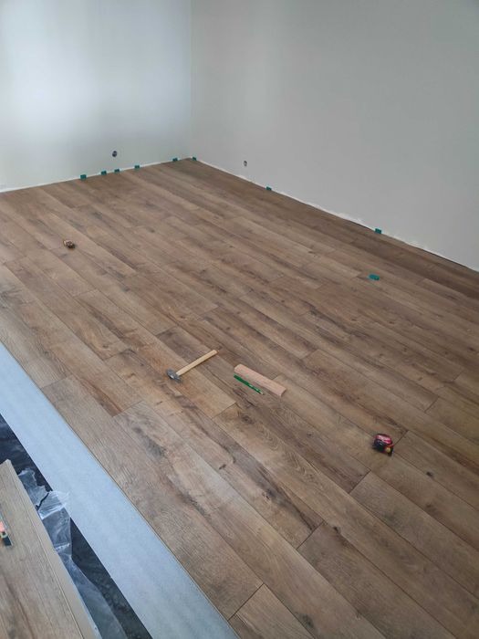 Układanie paneli podłogowych w jasnym pomieszczeniu, widoczne narzędzia: młotek, ołówek, miarka oraz kliny dystansowe przy ścianach.