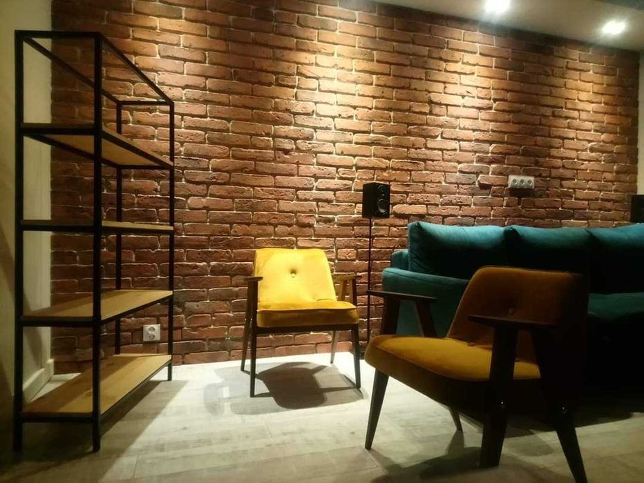 Salon z dekoracyjną ścianą z cegły, oświetlony punktowo, z metalowym regałem z drewnianymi półkami, dwoma fotelami w odcieniach żółci i turkusową kanapą.