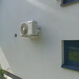 Biała jednostka zewnętrzna klimatyzatora marki Hyundai zamontowana na elewacji budynku z widocznym fragmentem okna i zwiniętym wężem ogrodowym w tle.