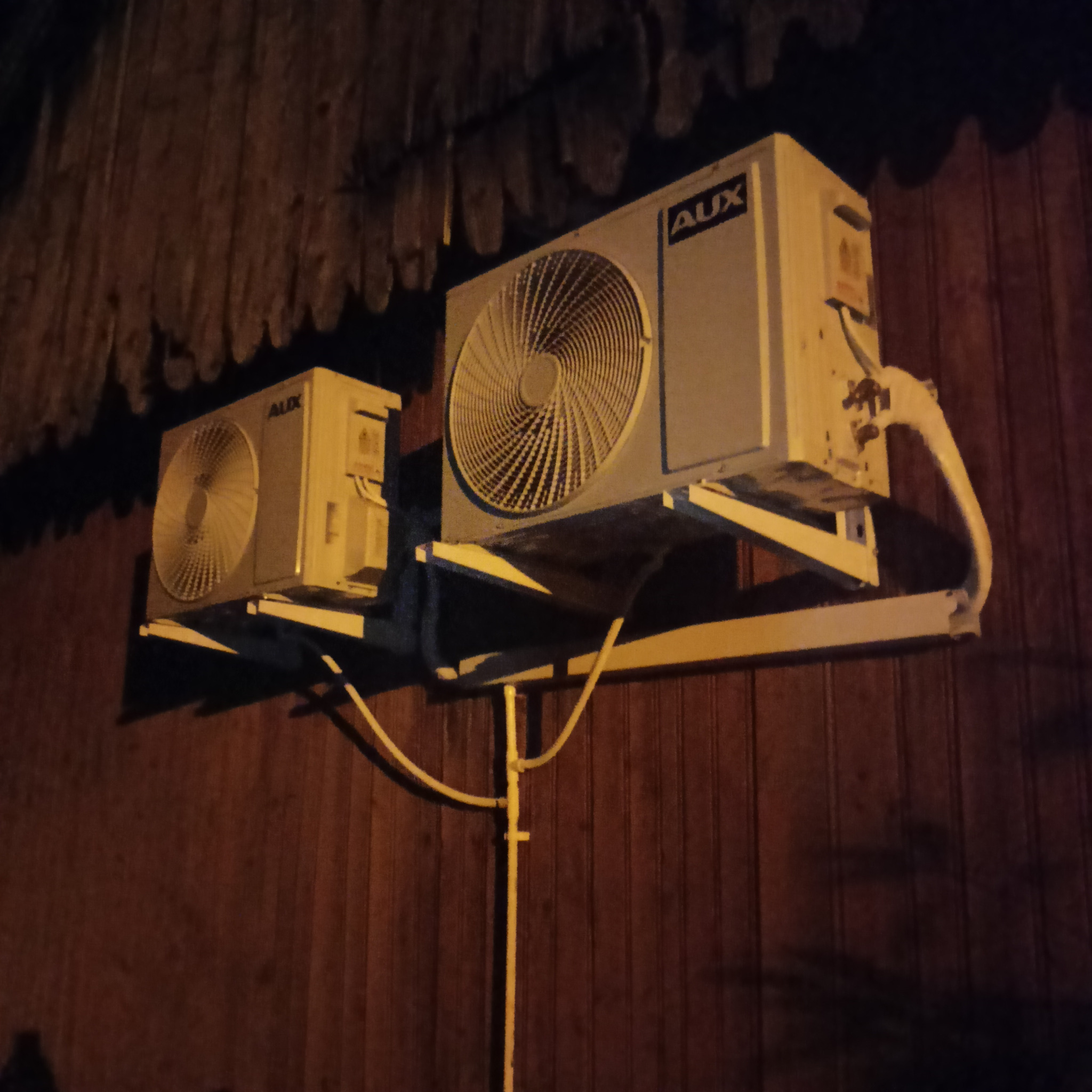 Dwa klimatyzatory marki AUX zamontowane na zewnątrz budynku z drewnianą elewacją, oświetlone ciepłym światłem, widoczne rury odprowadzające skropliny.