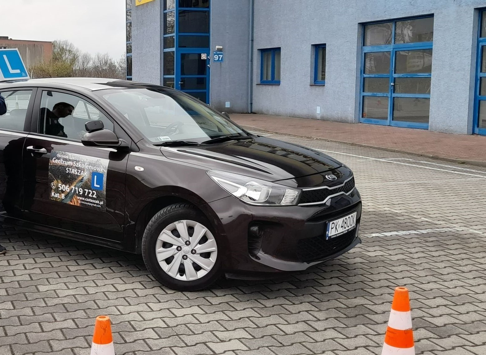 Brązowy samochód z naklejką 'L' i logo szkoły kierowców Centrum Szkoleniowe Staszak, zaparkowany na brukowanej powierzchni z pomarańczowymi pachołkami, budynek w tle.