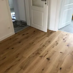 Ukończona podłoga z deski warstwowej z wyraźnymi sękami wypełnionymi czarną szpachlą, widoczna w przejściu między dwoma pomieszczeniami oddzielonymi białymi drzwiami.