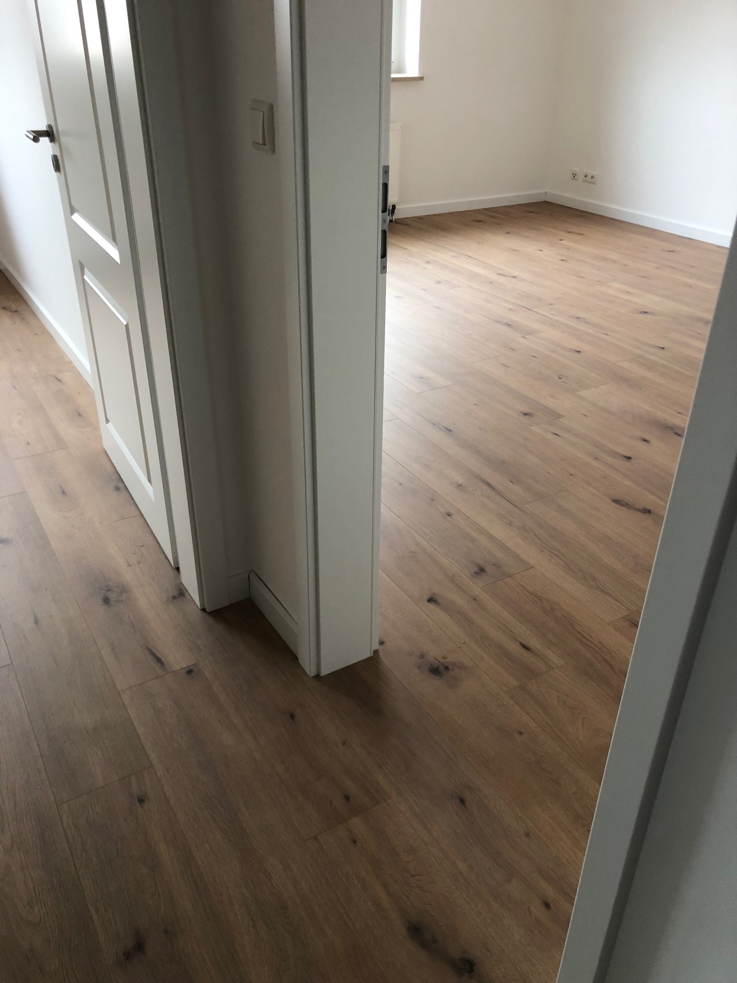 Widok na nowo ułożoną podłogę z paneli winylowych o wzorze drewna, z widocznymi białymi drzwiami i listwami przypodłogowymi, sfotografowaną z perspektywy wnętrza pokoju.