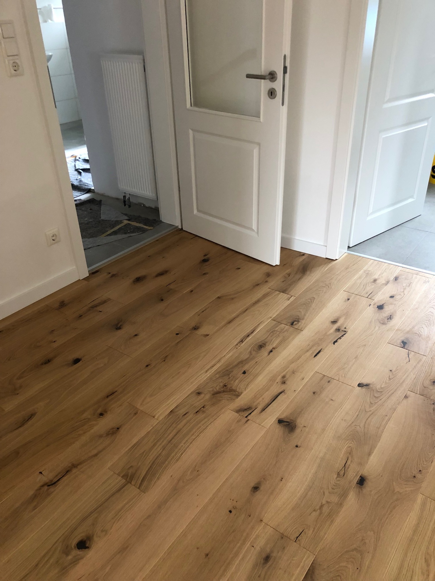 Ukończona podłoga z deski warstwowej z wyraźnymi sękami wypełnionymi czarną szpachlą, widoczna w przejściu między dwoma pomieszczeniami oddzielonymi białymi drzwiami.