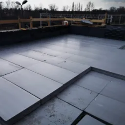 Układanie szarych płyt izolacyjnych na dachu budynku, widoczne czarne membrany izolacyjne pionowych ścianek, w tle konstrukcja stalowa i drzewa.